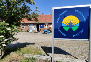 Willkommensschild am Ballum Campingplatz in Süd-Dänemark mit Gebäude und blühendem Baum.