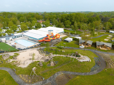 Vista aérea de un camping con tiendas safari, piscina, tobogán acuático y área de juegos en entorno verde.