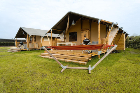 Tienda safari Villatent Nomad en Holiday Park Ackersate, Países Bajos, con terraza de madera y hamaca roja.