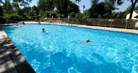 Piscina al aire libre con niños y adultos nadando y jugando, rodeada de árboles en un día soleado en Bélgica.