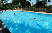 Udendørs swimmingpool med børn og voksne svømmende og legende, omgivet af træer på en solrig dag i Belgien.
