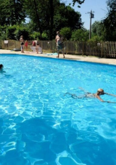 Piscina al aire libre con niños y adultos nadando y jugando, rodeada de árboles en un día soleado en Bélgica.