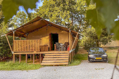 En safari telt med træveranda og parkeret bil ved siden af, omgivet af træer under åben himmel.