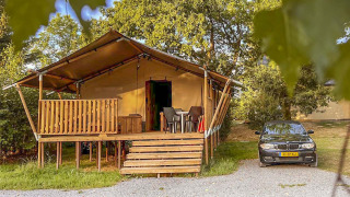 Safari tent with wooden veranda and parked car beside it, surrounded by trees in a natural setting.
