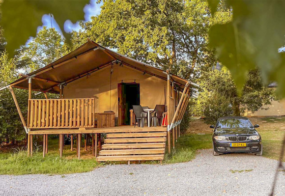 En safari telt med træveranda og parkeret bil ved siden af, omgivet af træer under åben himmel.