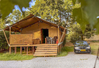 Safaritent met houten veranda en geparkeerde auto ernaast, omgeven door bomen in een natuurlijke omgeving.