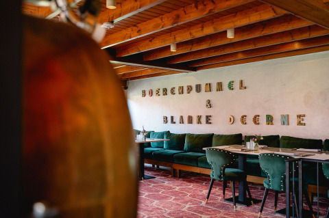 Interior moderno de restaurante con sillas verdes, techo de madera y 'BOERENDUMMEL & BLANKE DEERNE' en la pared.