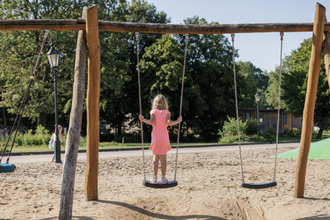 Una niña con vestido rosa está de pie sobre un columpio en Vakantiepark de Twee Bruggen, Gelderland, Países Bajos.