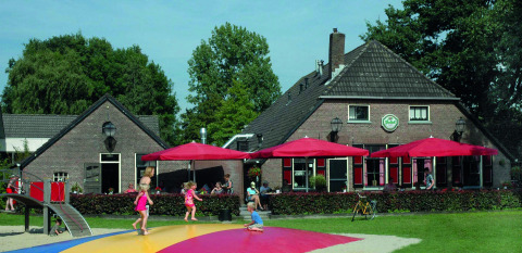 Niños juegan en parque de juegos junto a edificio de ladrillo y sombrillas rojas en Vakantiepark Sallandshoeve.