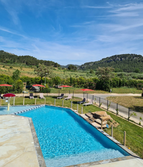 Piscina al aire libre con tumbonas y sombrillas rojas frente a colinas verdes en Camping De l'Olivigne, Occitanie.