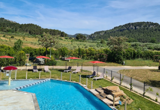 Swimmingpool med liggestole, røde parasoller og udsigt over grønne bakker på Camping De l'Olivigne i Occitanie.