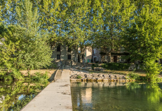 Pathway across a clear river at Camping De l'Olivigne in Occitanie, France, framed by lush green trees.