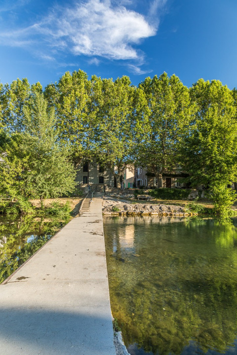 Passerella su un fiume limpido presso Camping De l'Olivigne in Occitania, Francia, con alberi verdi attorno.