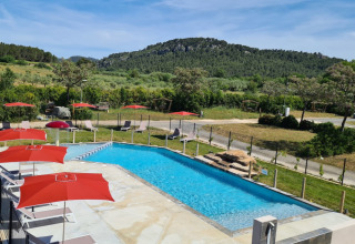 Piscina con ombrelloni rossi al Camping De l'Olivigne in Occitania, Francia, circondata da colline verdi.