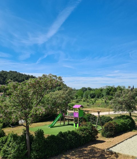 Parque infantil rodeado de vegetación bajo cielo azul en Camping De l'Olivigne, Occitania, Francia.