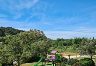 Legeplads omgivet af grønne træer under en blå himmel hos Camping De l'Olivigne i Occitanie, Frankrig.