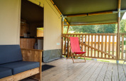 Tienda safari con servicios sanitarios, terraza de madera, silla roja y sofá, vista a la naturaleza.