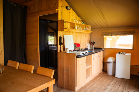 Coin cuisine dans une tente safari avec meubles en bois, réchaud à gaz, mini-frigo et fenêtre extérieure.