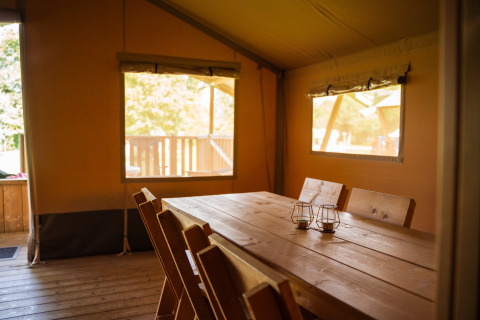 Interno di una tenda safari con tavolo e sedie di legno al Camping de Chênefleur in Belgio, illuminata dal sole.