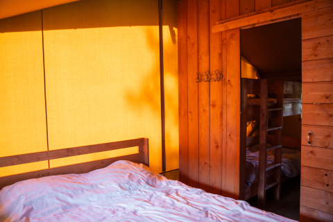 Vue intérieure d'une tente safari avec murs en bois, lit double et lits superposés éclairés chaudement.
