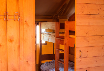 Vista interna di tenda safari con pareti in legno, letti a castello e servizi igienici in luce calda.