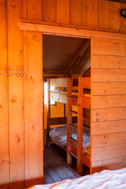 Interior view of a safari tent with wooden walls, bunk beds, and sanitary facilities in warm lighting.