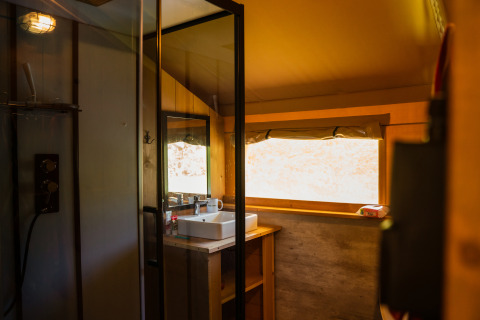 Intérieur d’une tente safari avec salle de bain privée, lavabo, grande fenêtre et éclairage moderne.