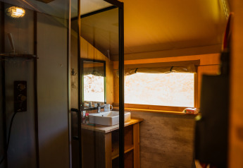 Interno di una tenda safari con bagno privato, lavandino, ampia finestra e illuminazione moderna.