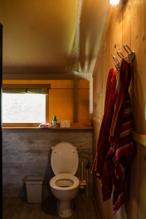 Interior de un baño de tienda safari con inodoro, ventana, toallas colgadas y luz cálida.