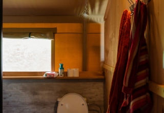 Interior de un baño de tienda safari con inodoro, ventana, toallas colgadas y luz cálida.