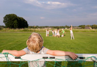 Ein Kind lehnt am Fußballtor, während andere Kinder auf dem Rasen bei FarmCamps De Smulhoeve spielen.