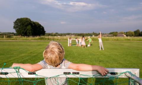 Ein Kind lehnt am Fußballtor, während andere Kinder auf dem Rasen bei FarmCamps De Smulhoeve spielen.