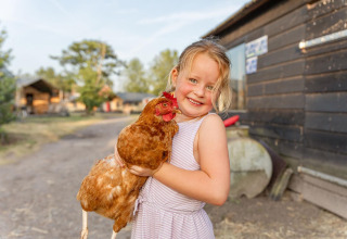 Una niña sonriente con vestido a rayas sostiene una gallina marrón en FarmCamps Breehees, Brabante Septentrional, Países Bajos.