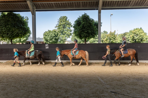 Børn rider på ponyer med ledsagere under et overdækket område hos FarmCamps Breehees i Nordbrabant.