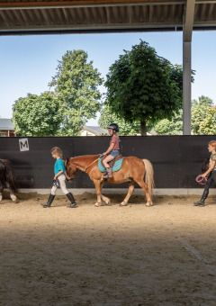 Niños montan ponis con guías en una pista cubierta en FarmCamps Breehees, Brabante Septentrional, Países Bajos.