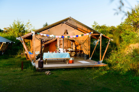 Safari tent with wooden deck, colorful bunting, and outdoor dining table set in lush green nature.