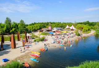 Alojamiento glamping con playa de arena, kayak y personas nadando junto a un lago rodeado de árboles verdes.