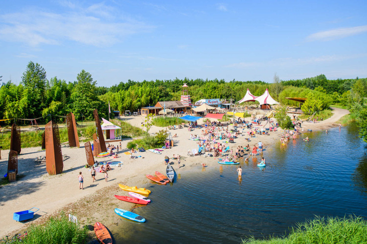 Glamping site with sandy beach, kayaks, people swimming and lounging by a lake, surrounded by green trees.