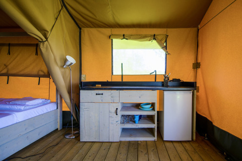 Innenraum eines Safarizeltes mit Küchenzeile, kleinem Kühlschrank, Tellerregal und Bett auf Holzboden.