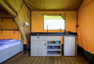 Safari tent interior featuring a small kitchenette, mini fridge, and bed, all set on a wooden floor.