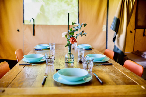 A rustic wooden dining table set for six inside a safari tent, featuring turquoise dishes and flowers.