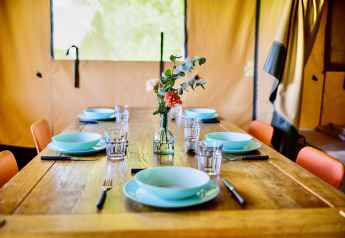 A rustic wooden dining table set for six inside a safari tent, featuring turquoise dishes and flowers.