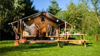 Safari tent set in lush greenery with a wooden picnic table outside, surrounded by trees and blue sky.