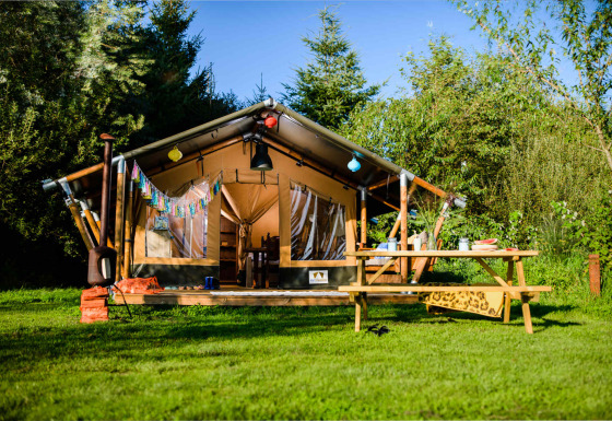 Tienda safari en un prado verde con mesa de picnic exterior, rodeada de árboles y cielo azul brillante.