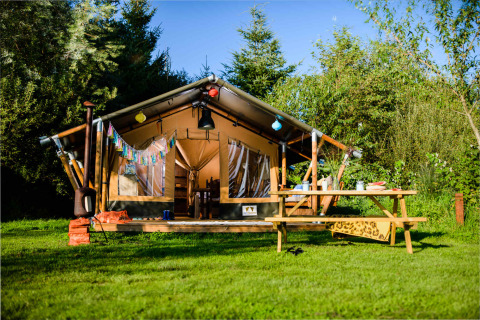 Safari tent set in lush greenery with a wooden picnic table outside, surrounded by trees and blue sky.