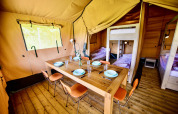 Interior of a safari tent featuring a wooden dining table, chairs, and cozy sleeping areas with beds.
