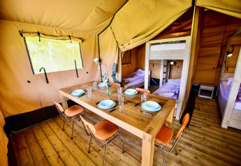 Het interieur van een safaritent met houten vloer, eettafel, oranje stoelen en slaapkamers met bedden.