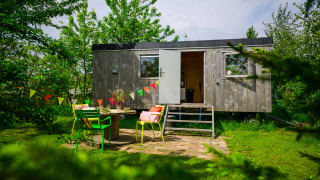 Encantadora tiny house Pipowagen con puerta abierta, sillas coloridas y banderines en un jardín verde.