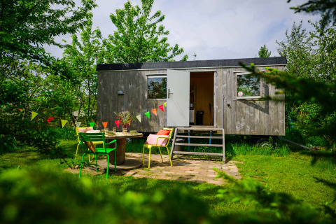 Charmante tiny house Pipowagen avec porte ouverte, chaises colorées et fanions dans un jardin verdoyant.