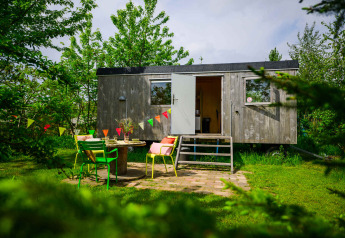 Gemütliches Pipowagen Tiny House mit geöffneter Tür, bunten Stühlen und Wimpeln im grünen Garten.